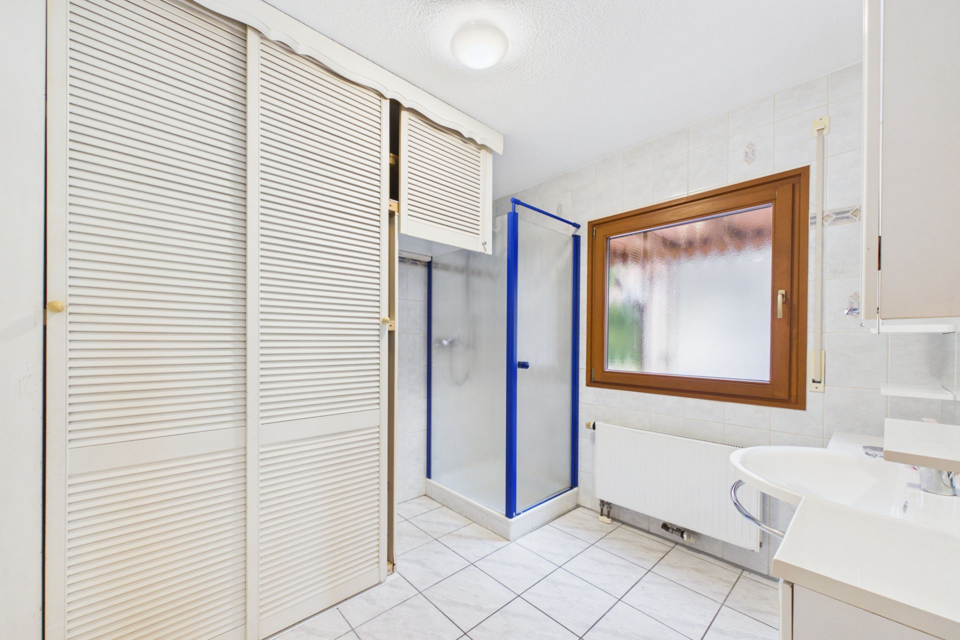 Helles Bad mit Dusche, großem Fenster, Waschbecken und Einbauschrank mit Lamellentüren.