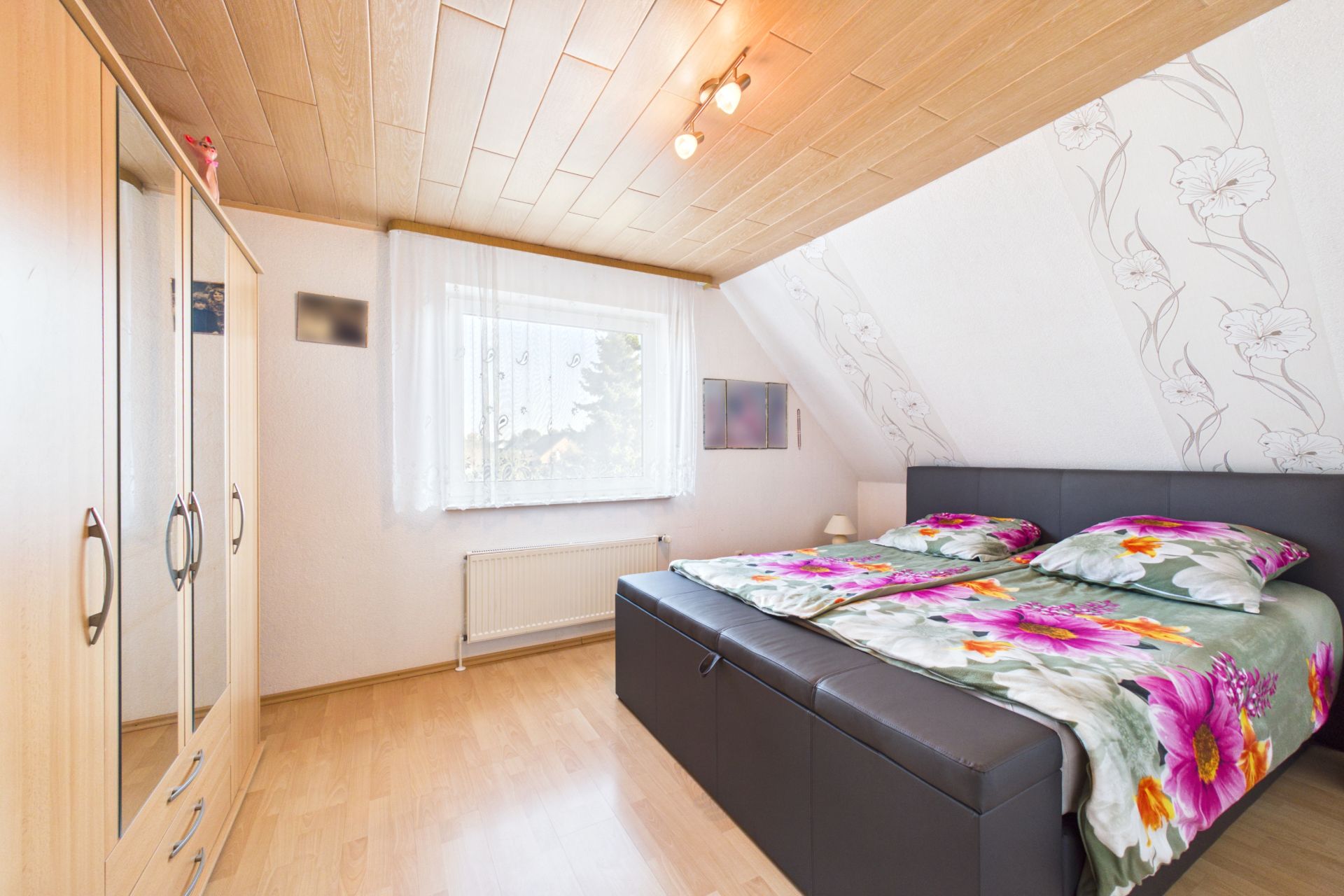 Helles Schlafzimmer mit Doppelbett, Blumenbettwäsche, Kleiderschrank und Fenster mit weißen Vorhängen.