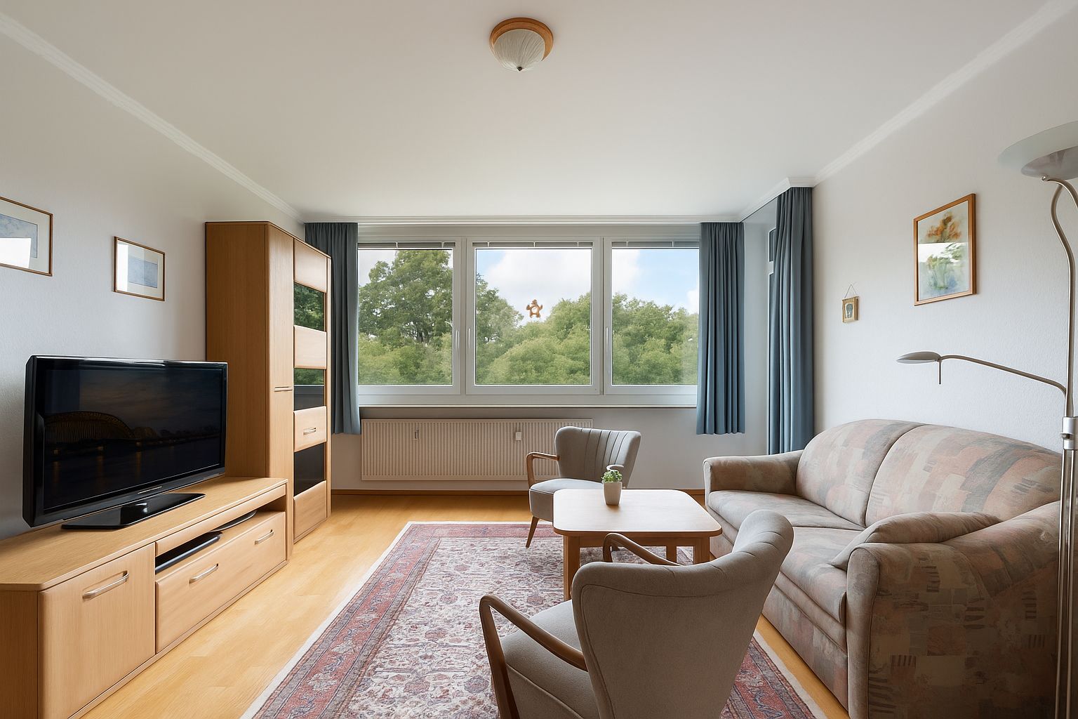Helles Wohnzimmer mit Sofa, zwei Sesseln, Teppich, Fernseher und großen Fenstern mit Blick ins Grüne.