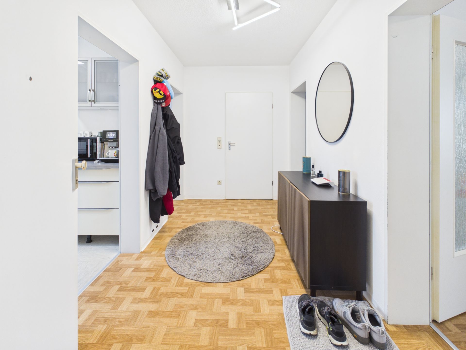 Heller Flur mit Parkettboden, rundem Teppich, Garderobe, Schuhen und Spiegel über Kommode.