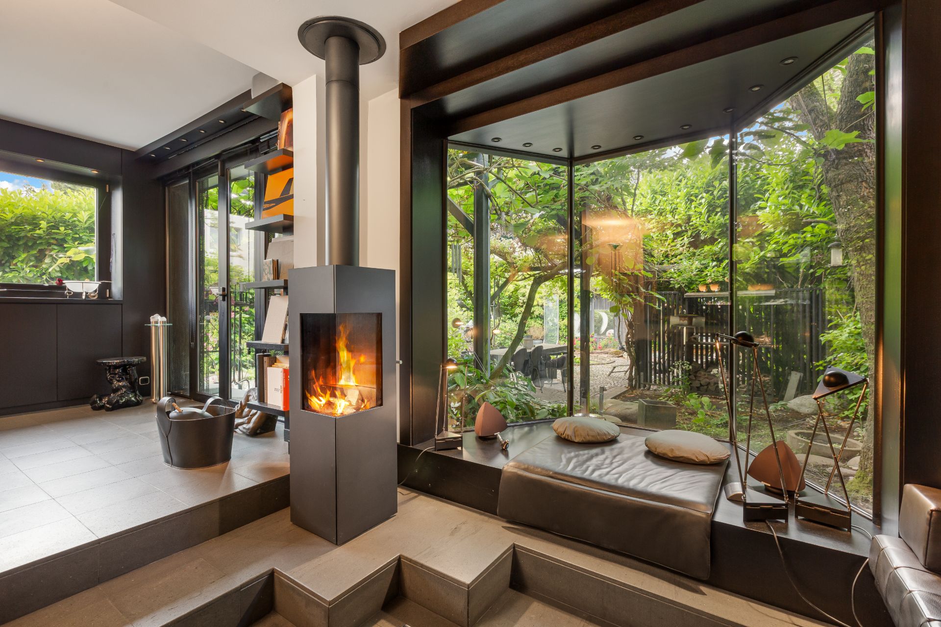 Modernes Wohnzimmer mit Kamin, großen Fenstern und gemütlicher Sitzbank mit Kissen vor Gartenblick.