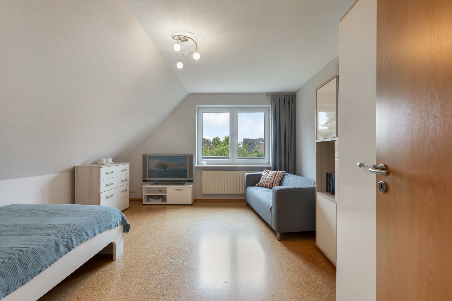 Helles Zimmer mit Bett, Sofa, Kommode und Fernseher, Fenster mit Blick nach draußen.