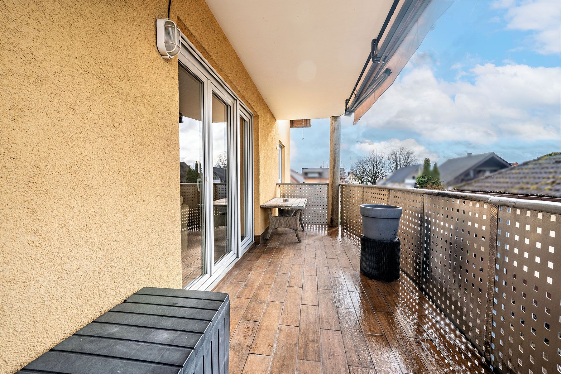 Überdachter Balkon mit Holzboden, Glastür, Tisch, Pflanztopf und Metallgeländer.