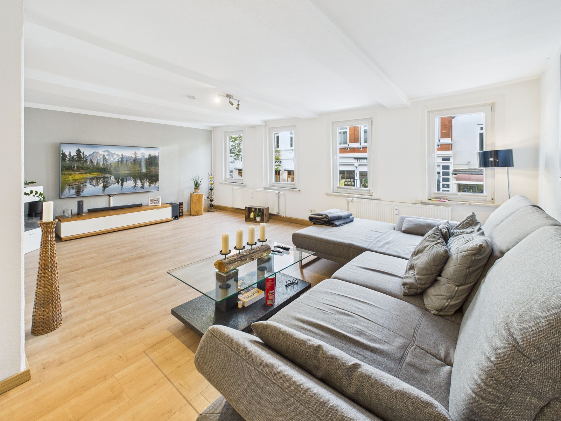 Modernes Wohnzimmer mit grauem Sofa, Glastisch und TV, helle Holzböden und drei Fenster.