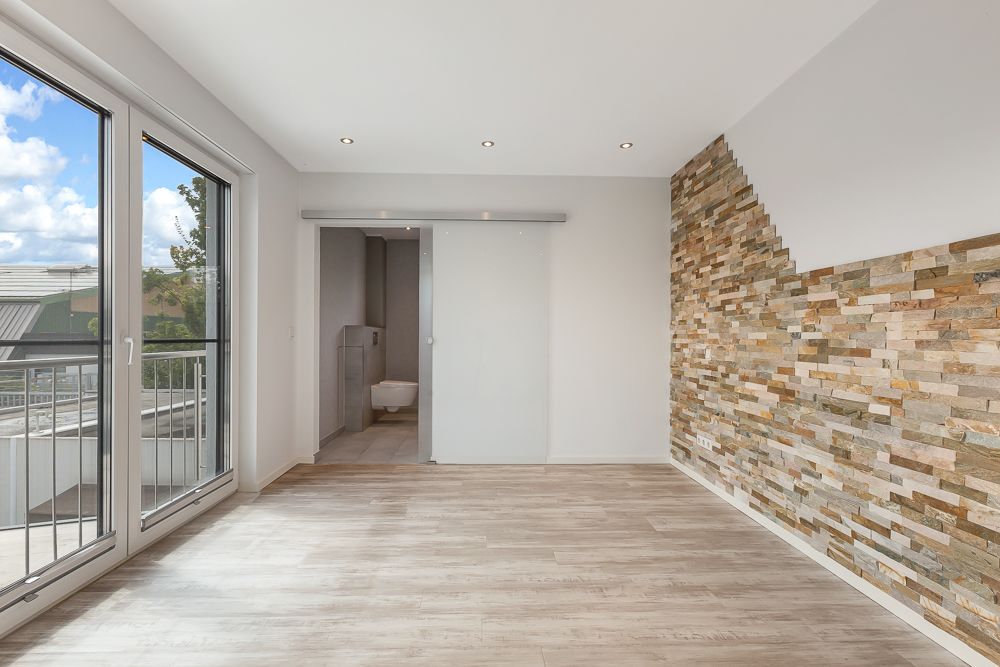 Helles Zimmer mit Steinwand, großer Fensterfront, Holzboden und Blick ins Bad mit Schiebetür.