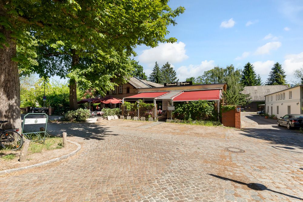 Gepflasterter Platz mit Bäumen und Gebäude mit roter Markise, Fahrrad links im Bild.