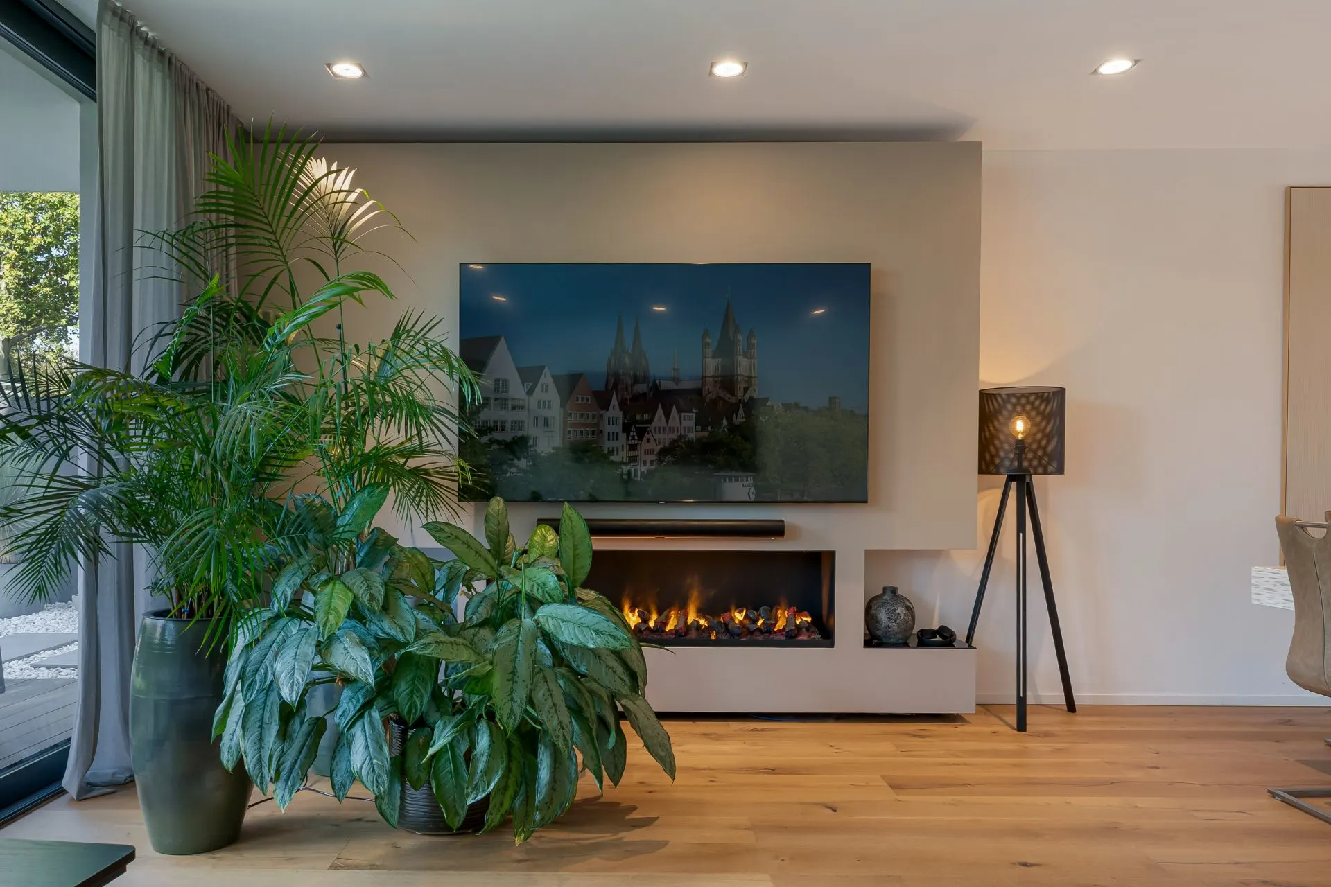 Wohnzimmer mit großem Fernseher, Kamin, mehreren Topfpflanzen und Stehlampe auf Holzfußboden.