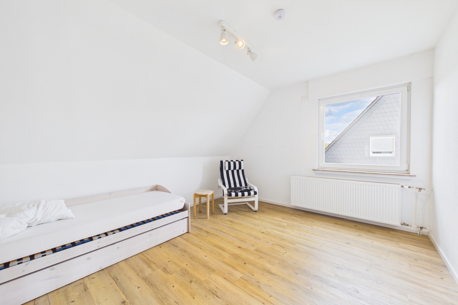 Helles Zimmer mit Einzelbett, kleinem Tisch, gestreiftem Sessel und Fenster mit Heizung.