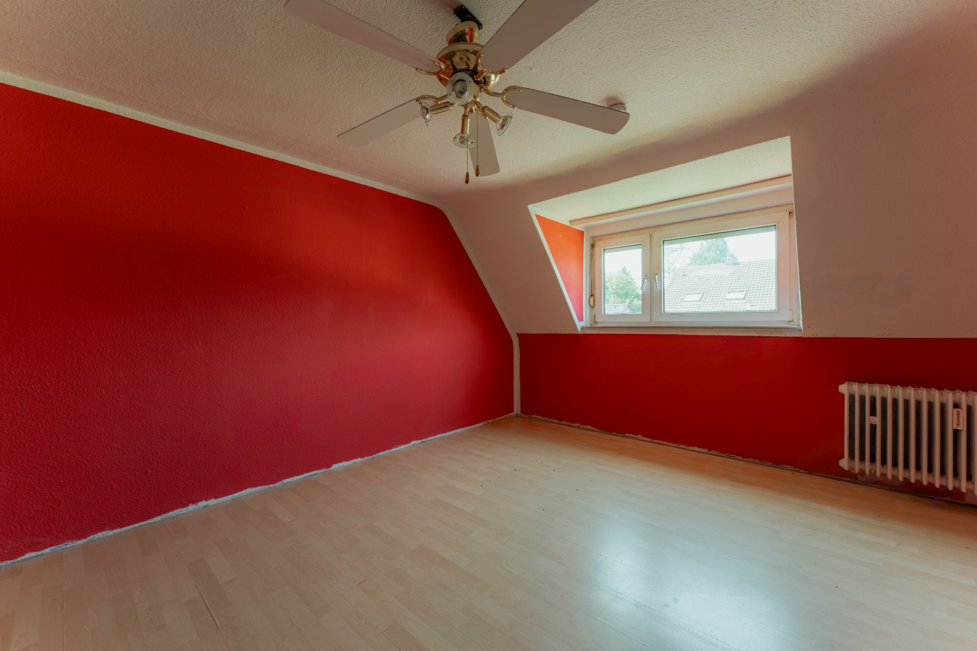 Leeres Zimmer mit roter Wand, Lamellenheizung, großem Fenster und Deckenventilator.