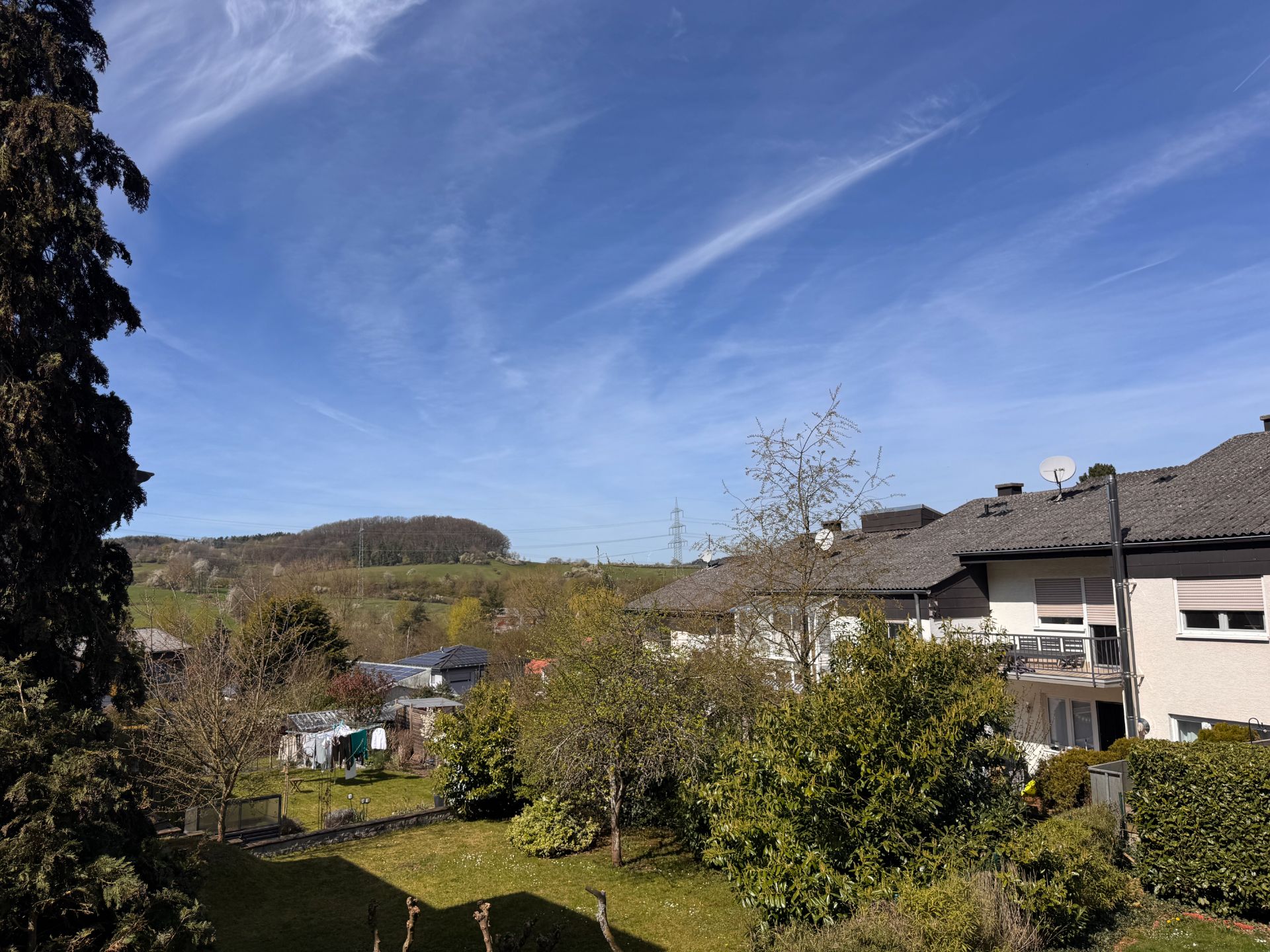 Blick auf ein Wohnhaus mit Garten, Bäumen und Wiese unter blauem Himmel.