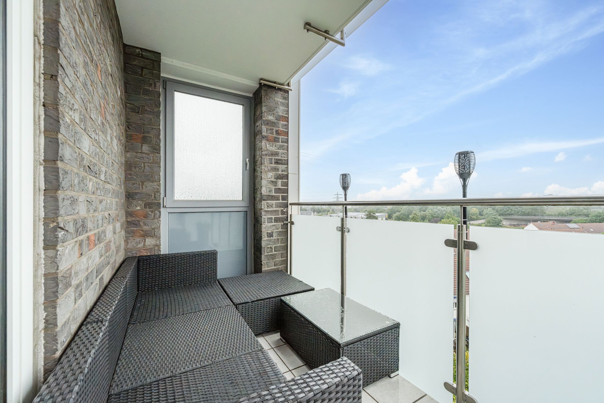 Balkon mit Rattanmöbeln, Glastisch und Ausblick auf Grünflächen unter blauem Himmel.