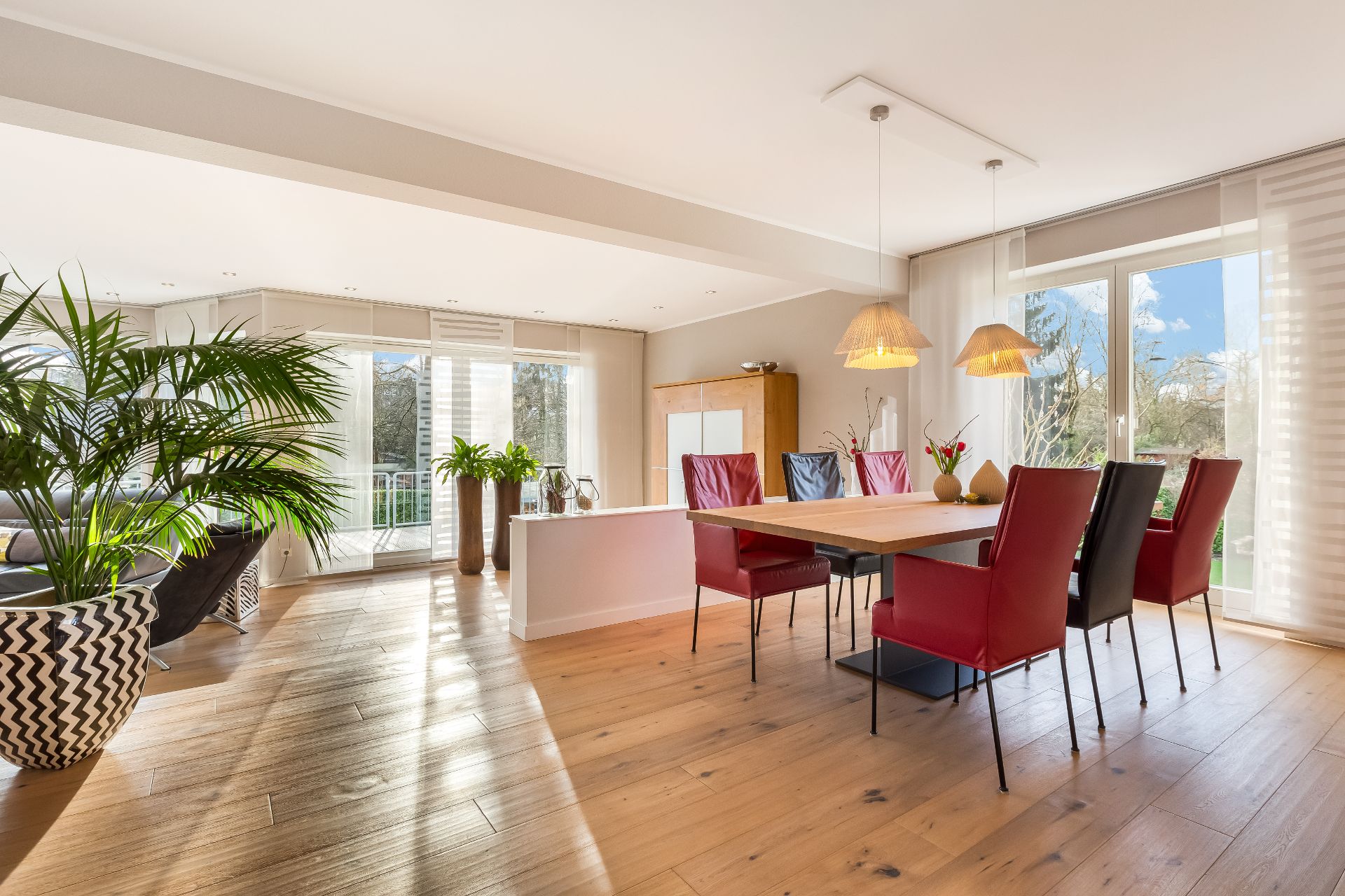 Helles Wohnzimmer mit Esstisch, roten Stühlen, großen Fenstern und Pflanzen.