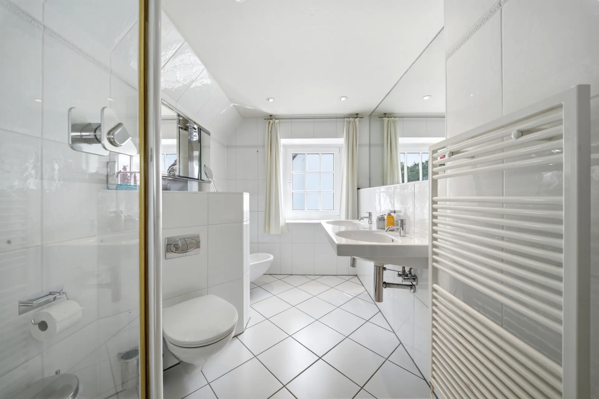 Helles Badezimmer mit WC, Waschbecken, Dusche, Heizkörper und zwei Fenstern.