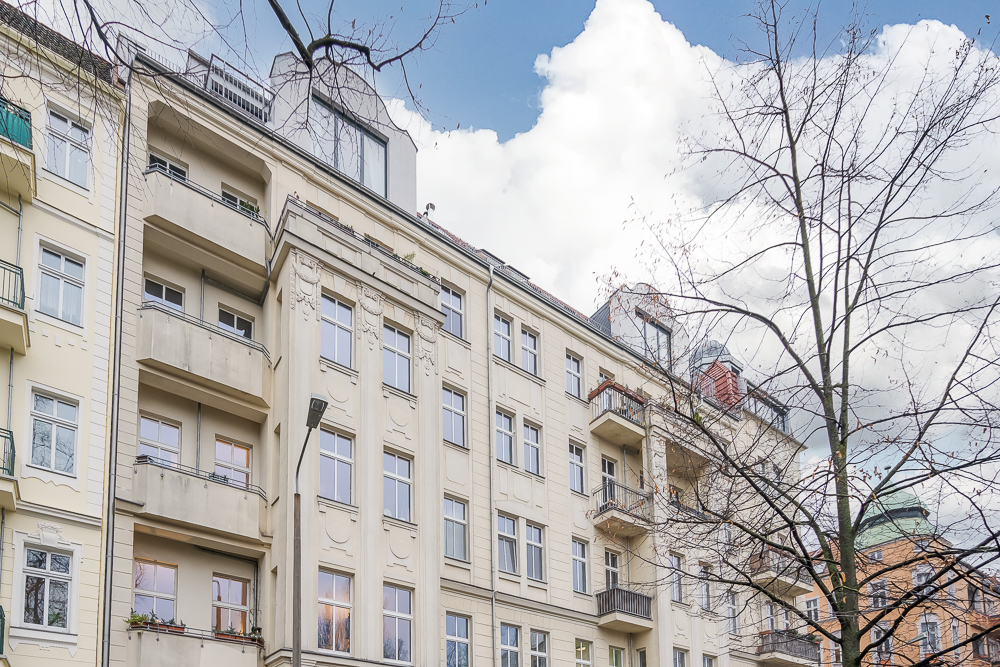 Mehrstöckiges Wohngebäude mit Balkonen und großen Fenstern, davor ein kahler Baum.