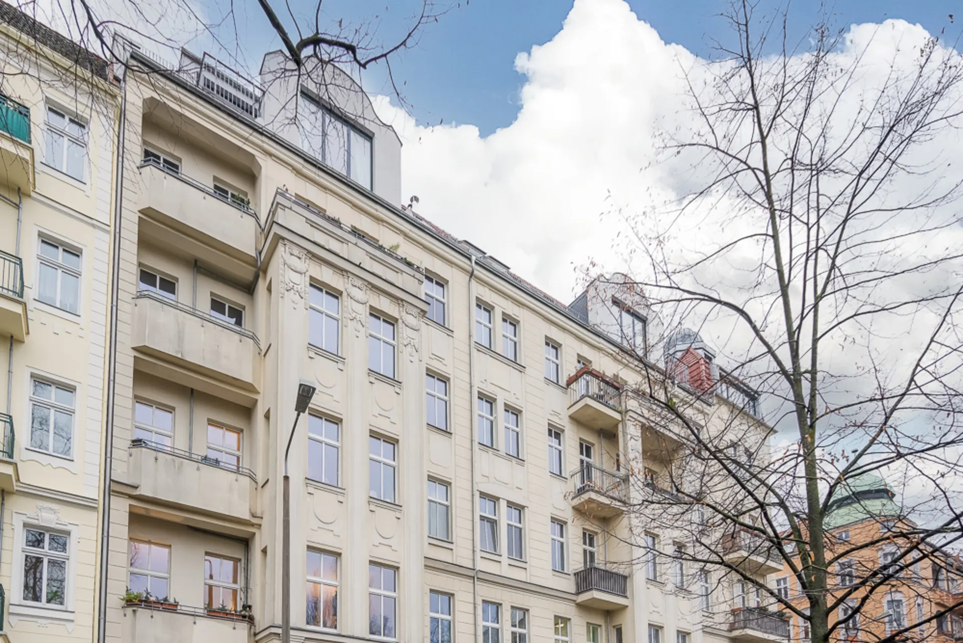 Mehrstöckiges Wohngebäude mit Balkonen und großen Fenstern, davor ein kahler Baum.
