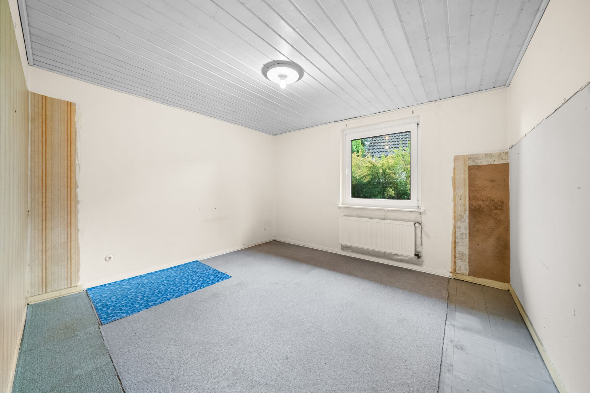 Leeres Zimmer mit Teppichboden, einem Fenster, weißer Decke und einem Heizkörper unter dem Fenster.
