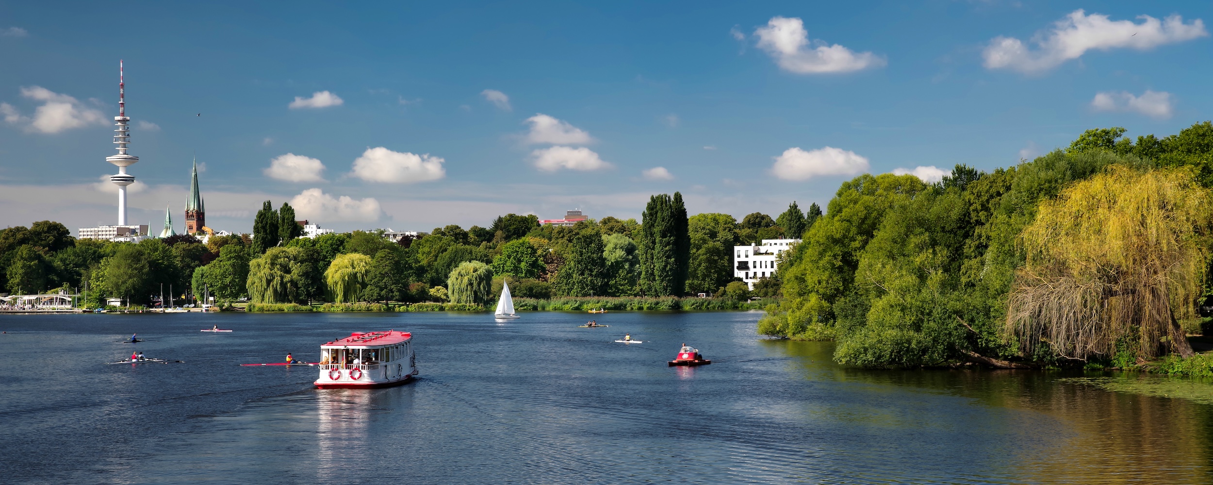 Fluss mit Booten und grüner Uferlandschaft, Häuser im Hintergrund, blauer Himmel.