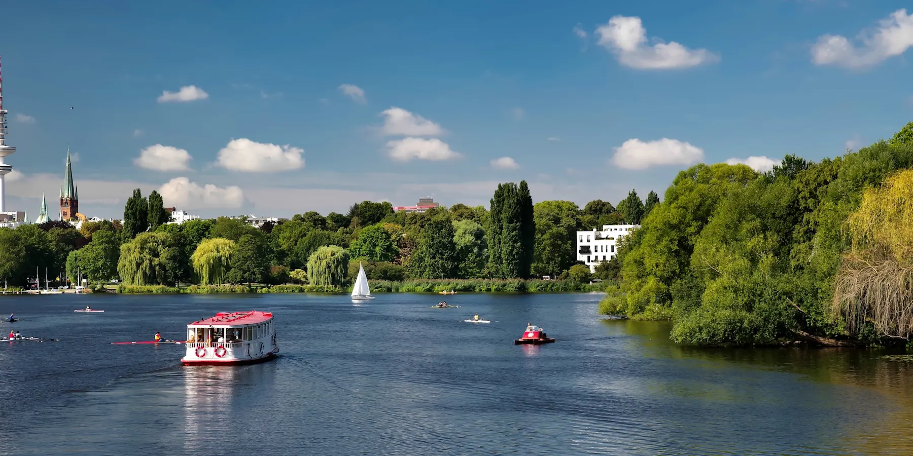 Fluss mit Booten und grüner Uferlandschaft, Häuser im Hintergrund, blauer Himmel.