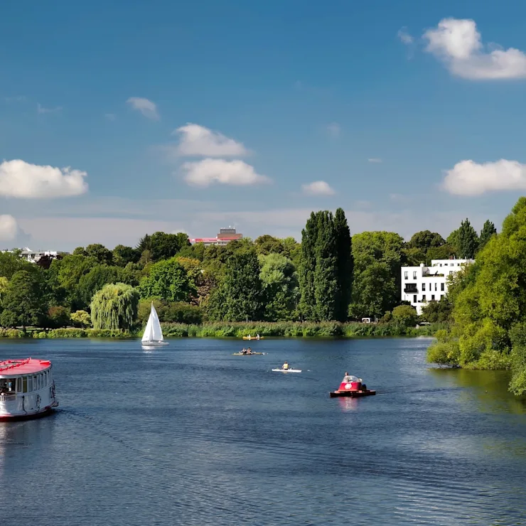 Fluss mit Booten und grüner Uferlandschaft, Häuser im Hintergrund, blauer Himmel.
