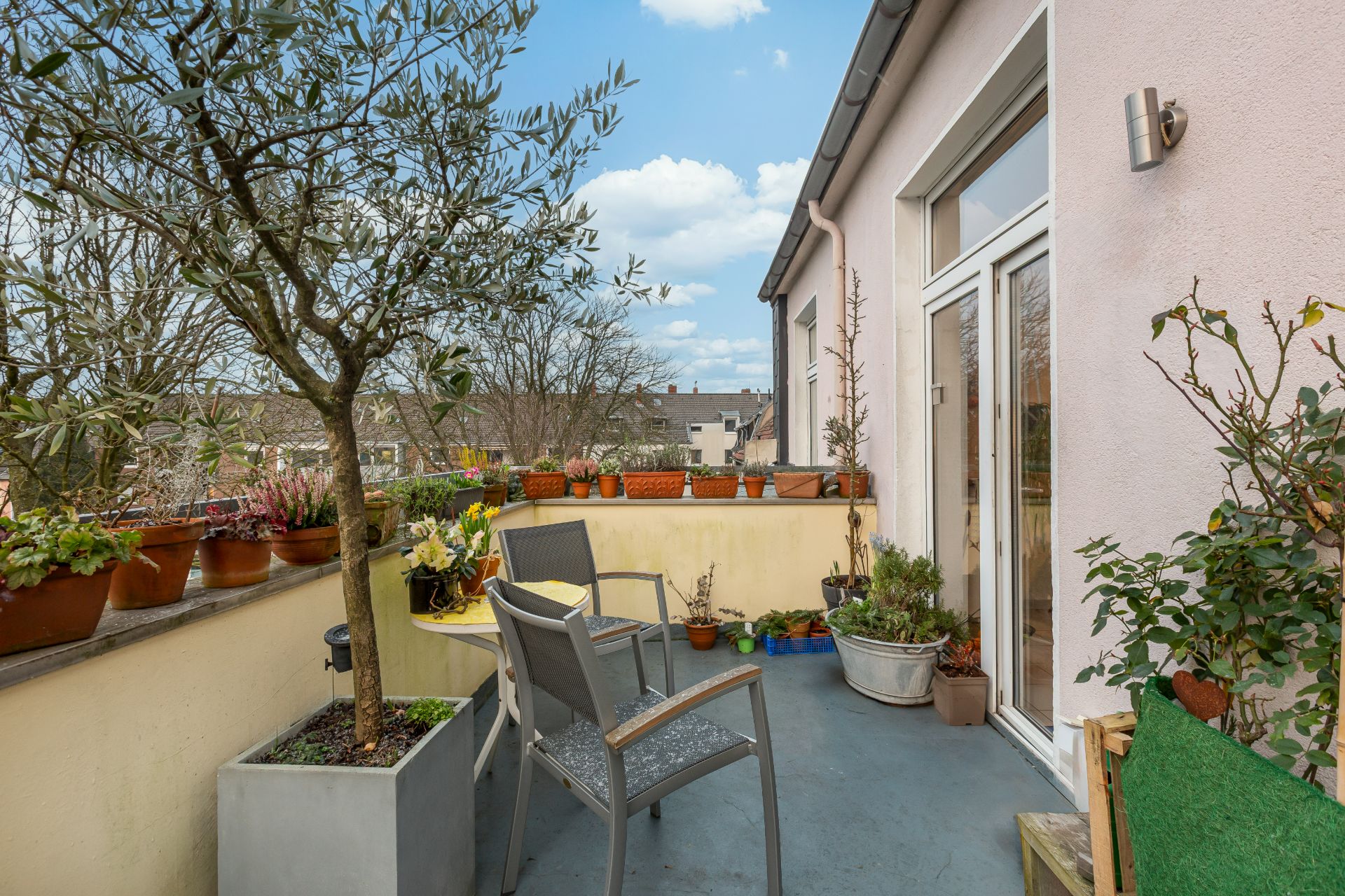 Balkon mit Pflanzen, Stühlen und Tisch, umgeben von Töpfen, mit Blick auf Nachbarhäuser.