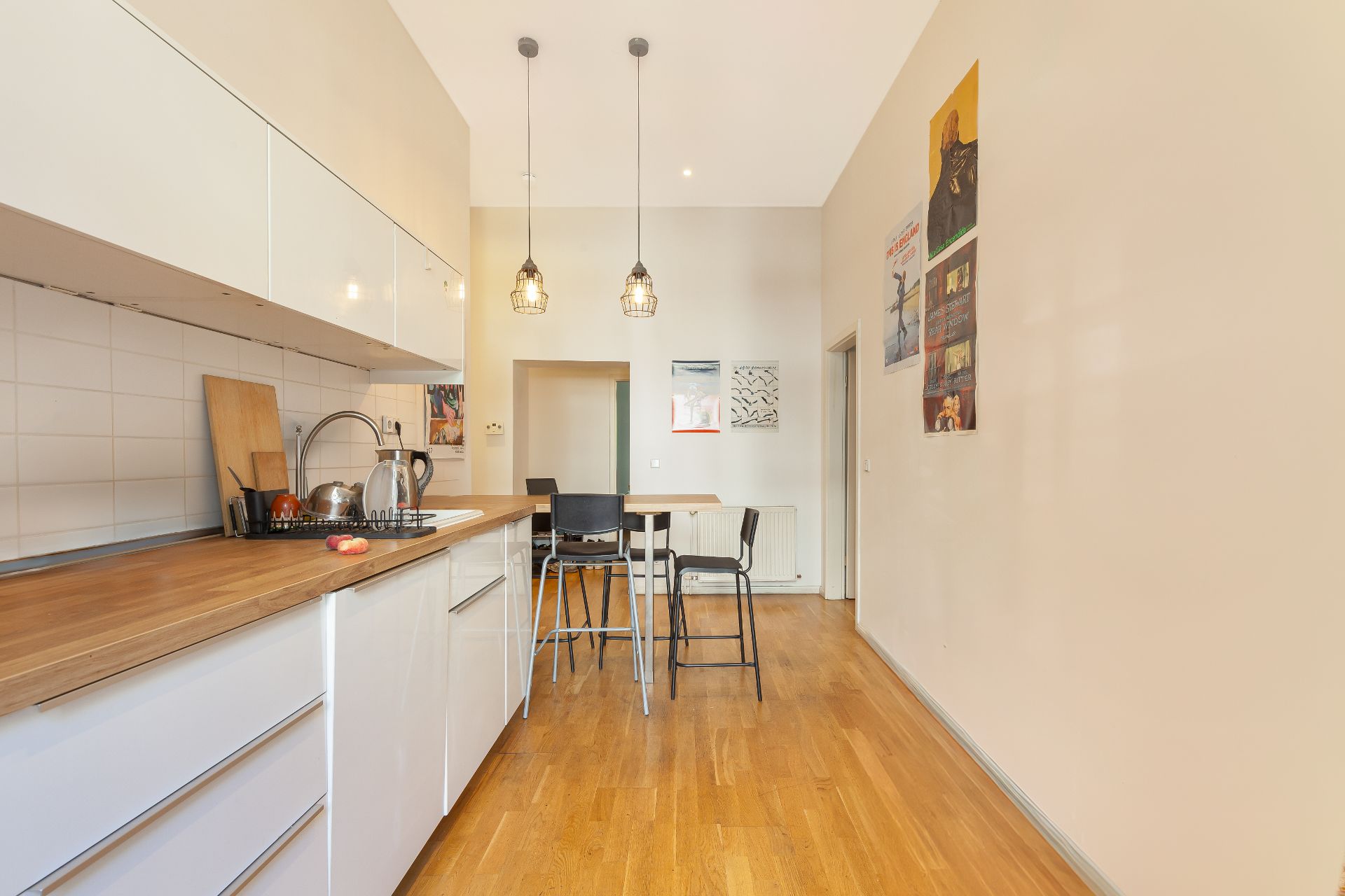 Moderne Küche mit Holzarbeitsplatte, Esstisch und Hockern, helle Wände, Holzboden, Wandbilder.