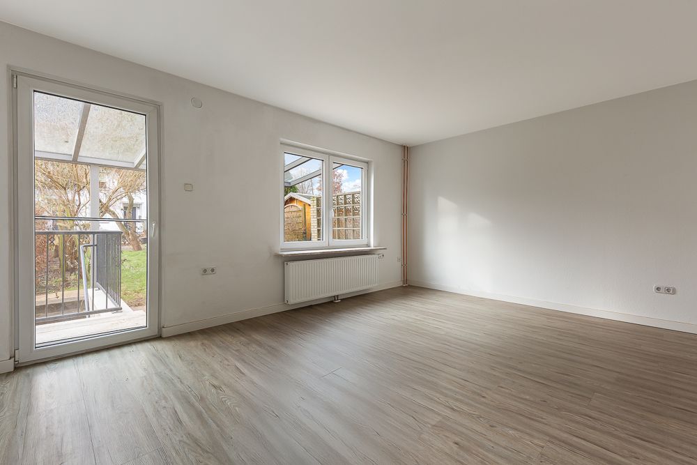 Helles, leeres Zimmer mit Fenster, Tür zur Terrasse und Holzfußboden.