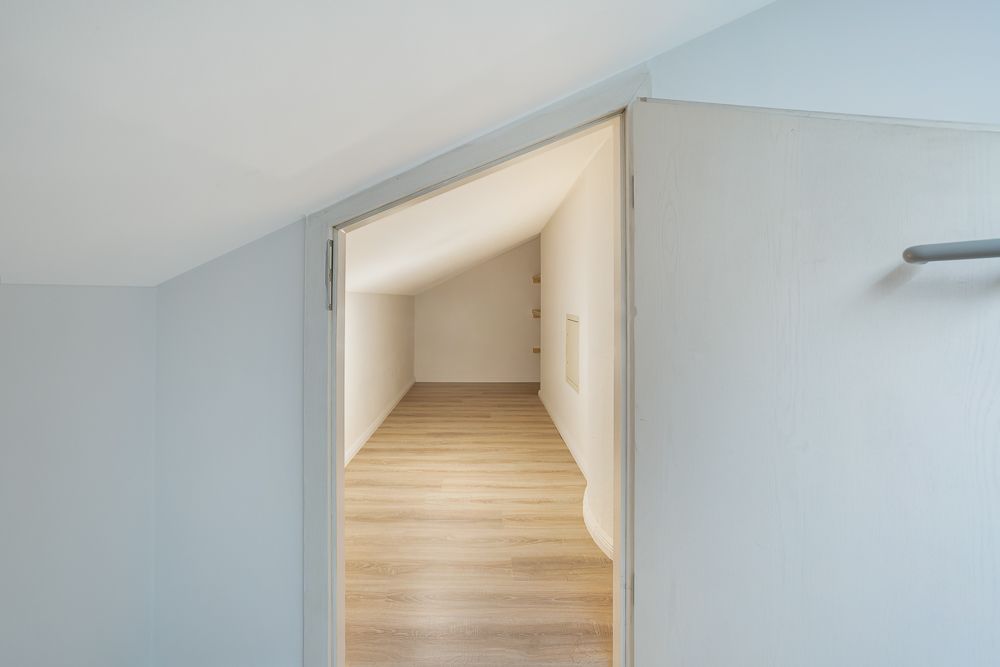 Blick durch eine Tür in einen kleinen, leeren Dachraum mit Holzfußboden.