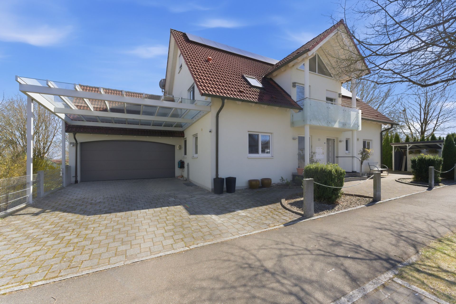 Modernes Einfamilienhaus mit rotem Dach, Glasüberdachung und Doppelgarage, davor gepflasterter Hof.