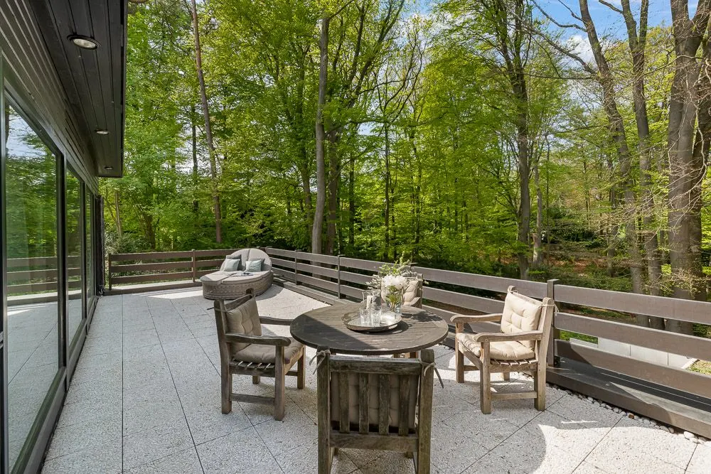 Terrasse mit rundem Tisch, drei Stühlen und einem Grill, umgeben von Bäumen.