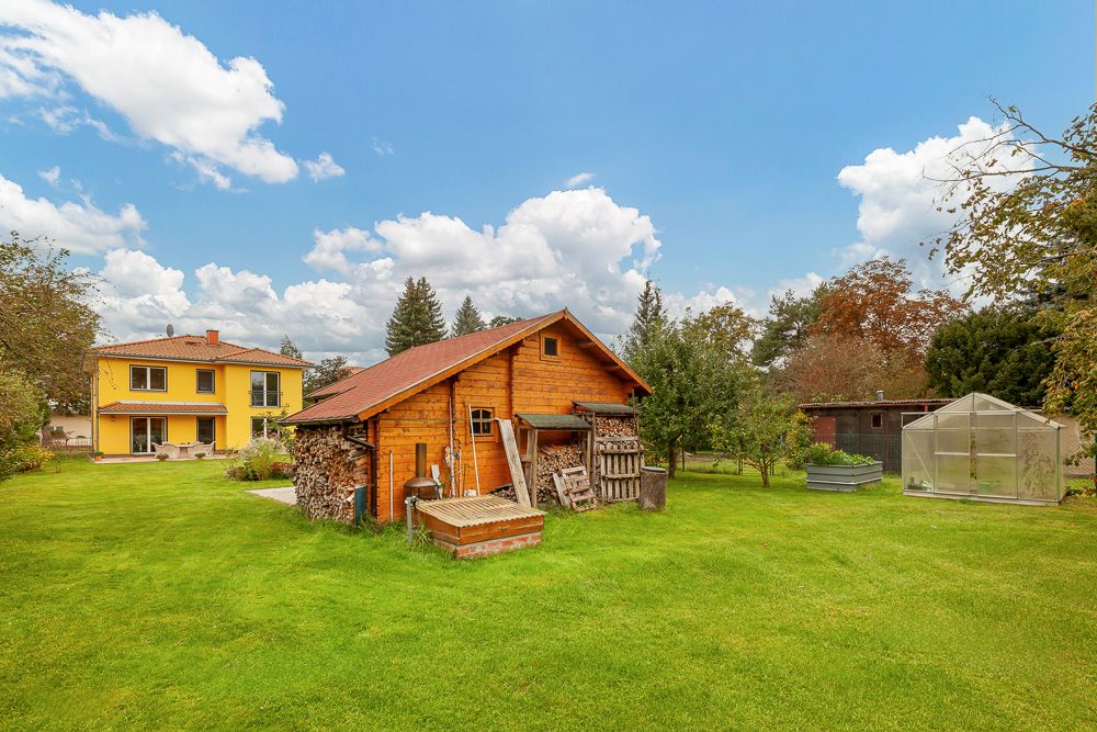 Großer Garten mit gelbem Haus, Holzschuppen und Gewächshaus bei blauem Himmel.