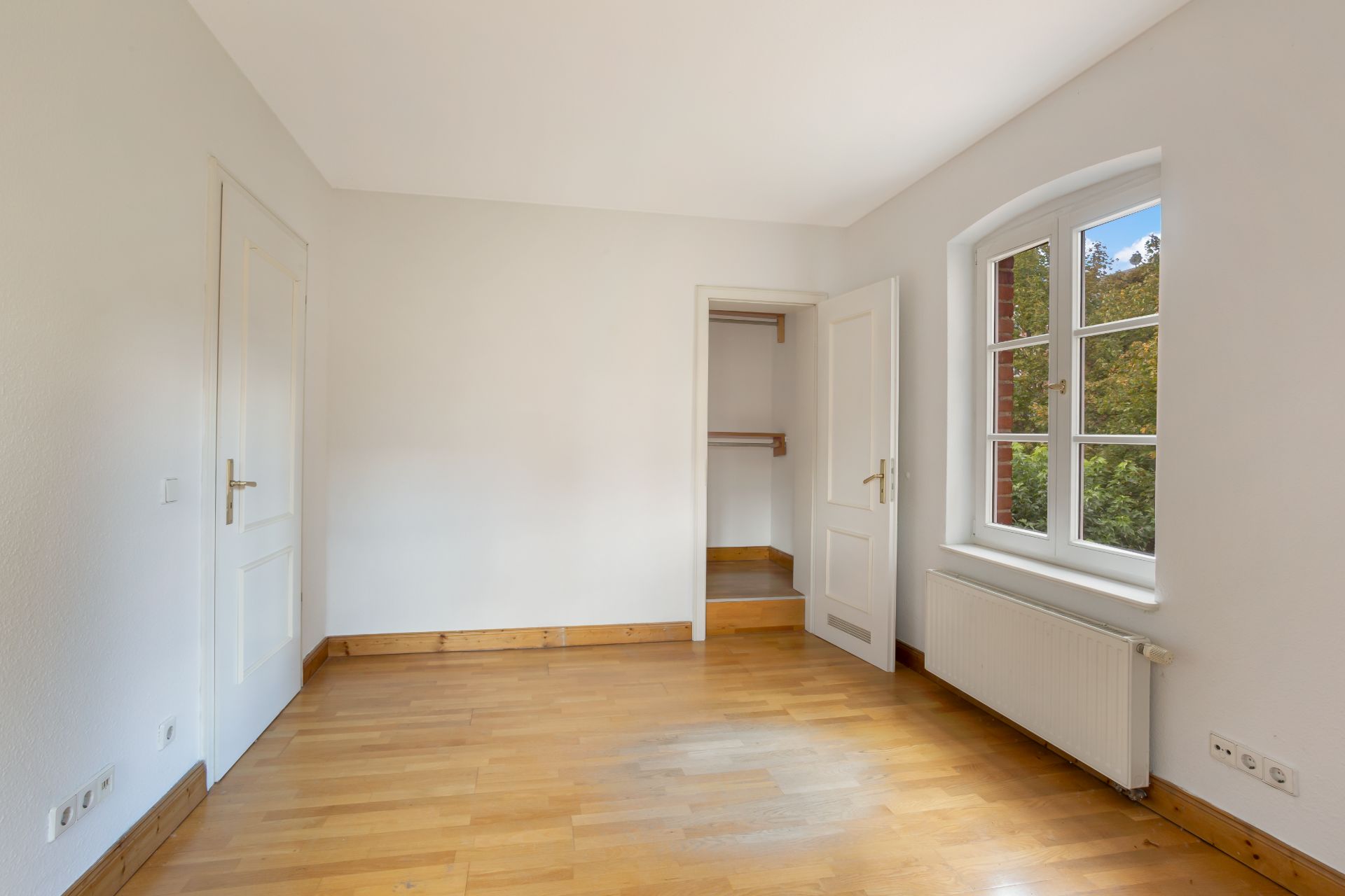 Leeres Zimmer mit Holzfußboden, weißer Wand, Fenster, Heizkörper und offenem Türbereich zu einem kleinen Raum.