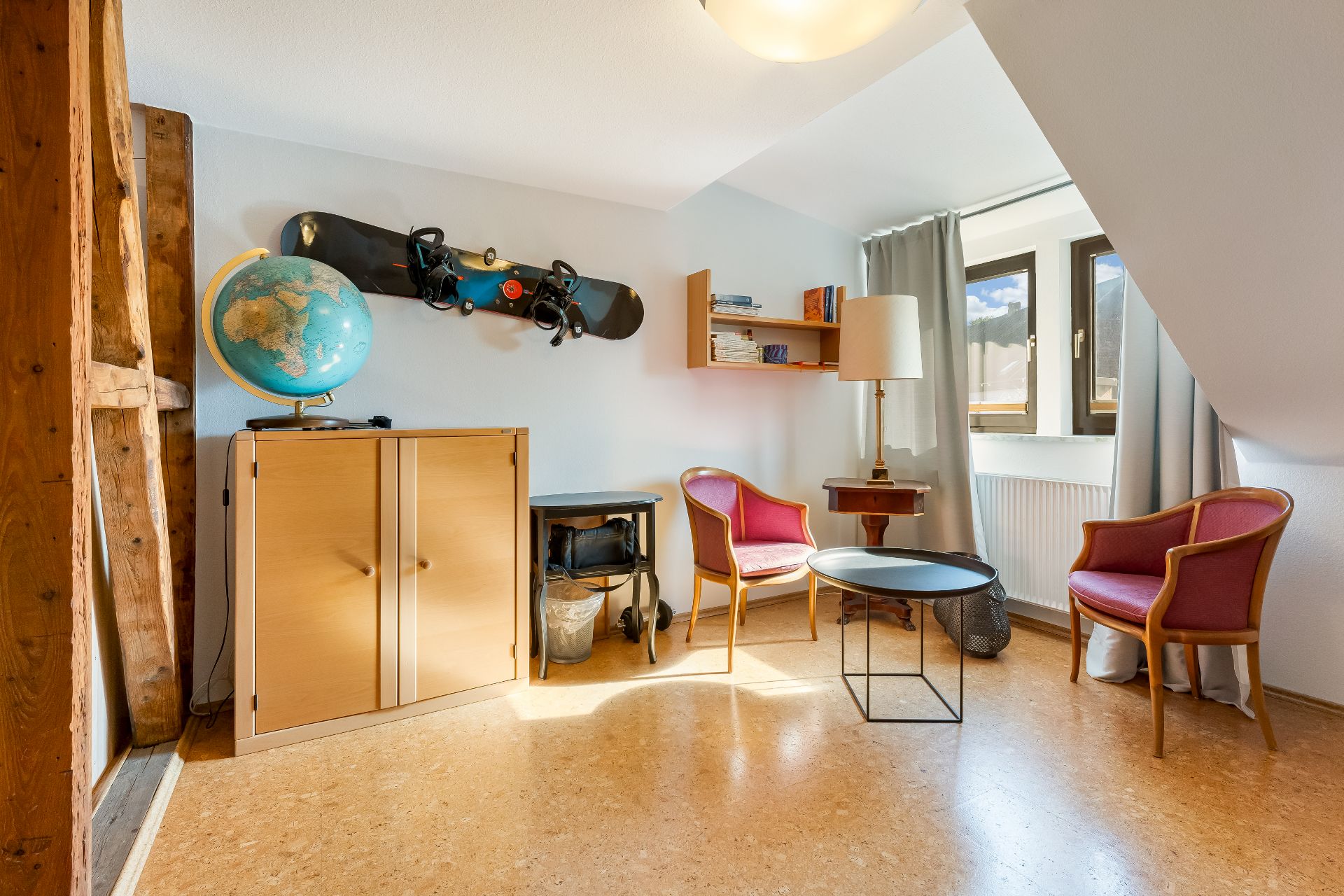 Helles Zimmer mit zwei Stühlen, rundem Tisch, Schrank, Globus und Snowboard an der Wand.