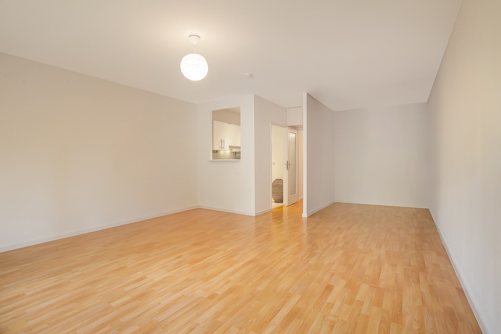 Helles, leeres Zimmer mit Holzfußboden und weiß gestrichenen Wänden, offene Küche sichtbar.