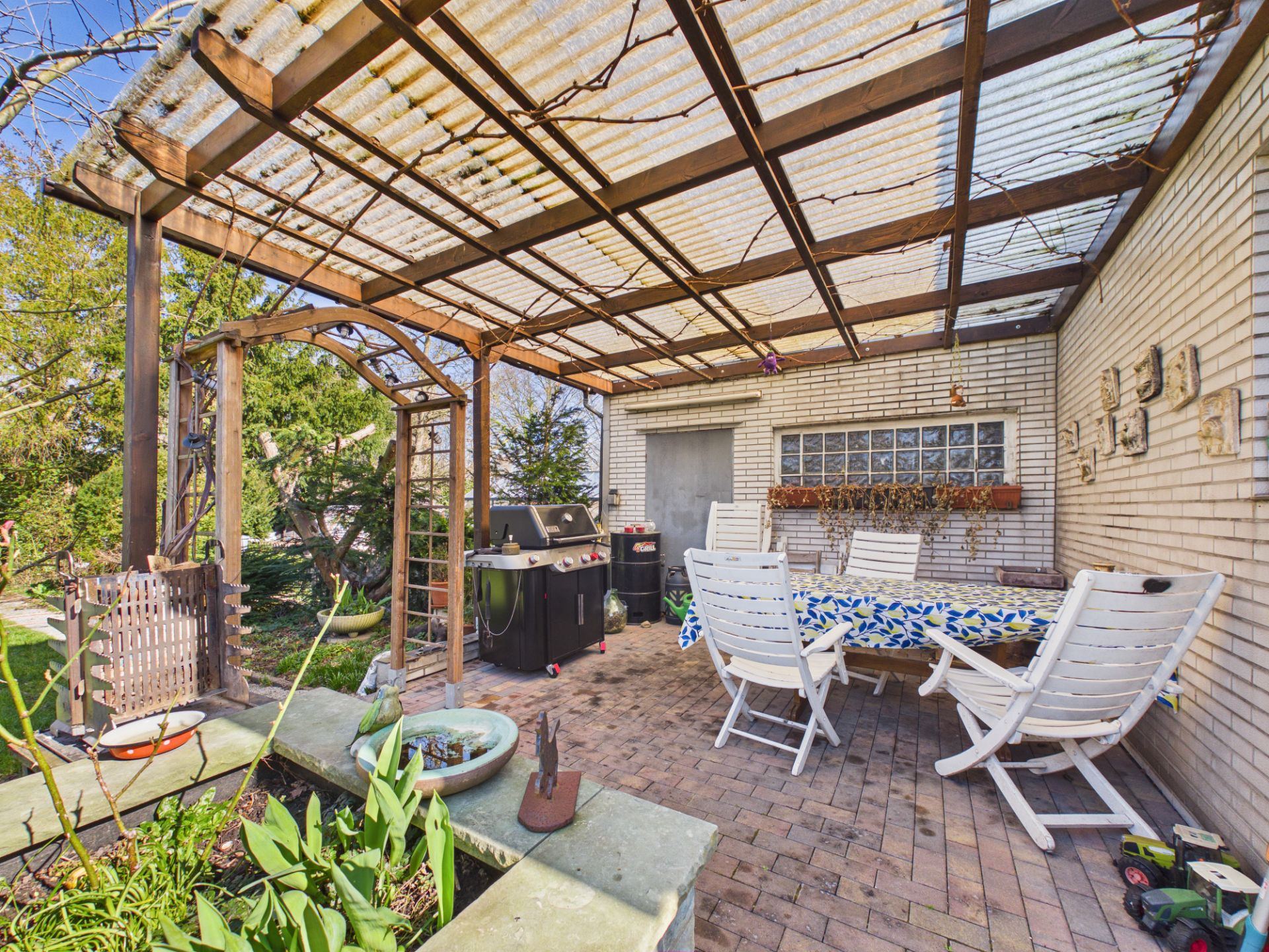 Überdachte Terrasse mit weißem Gartentisch, Stühlen und Grill neben einem Haus.