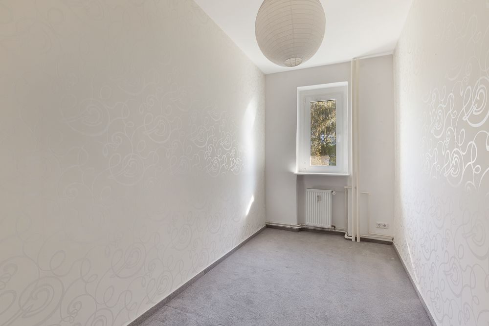 Schmaler, leerer Raum mit hellem Teppich, Fenster, Heizkörper und runder Deckenleuchte.