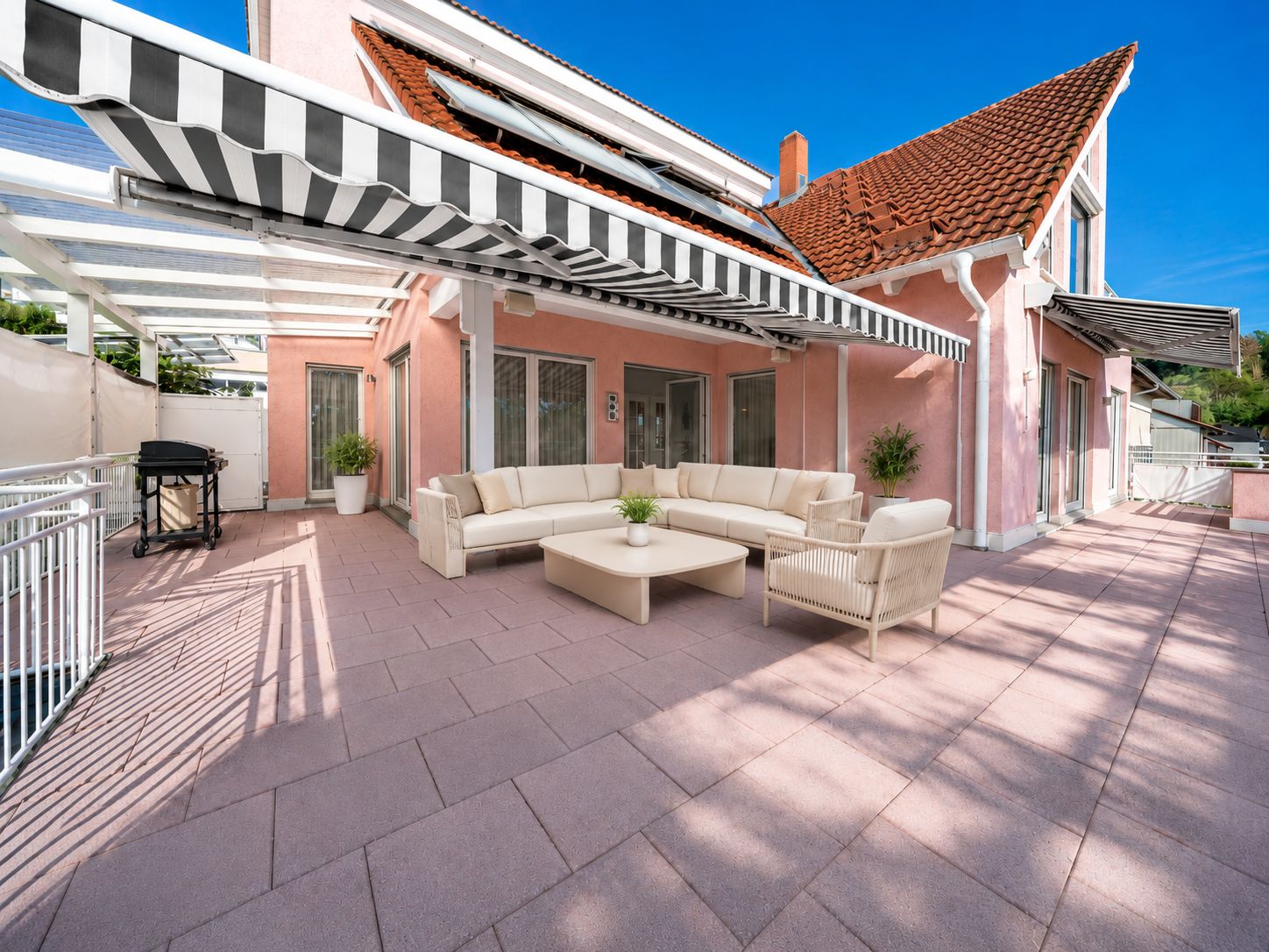 Große Terrasse mit weißen Gartenmöbeln, Markise und Grill an einem rosa Haus.