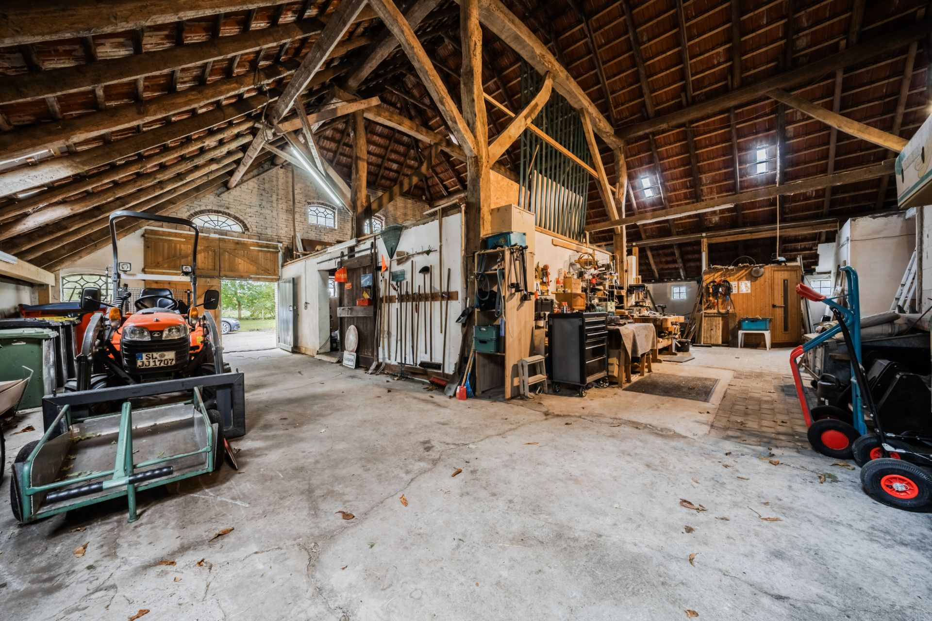 Große Scheune mit Traktor, Gartengeräten und Werkstattbereich, Holzgebälk sichtbar.