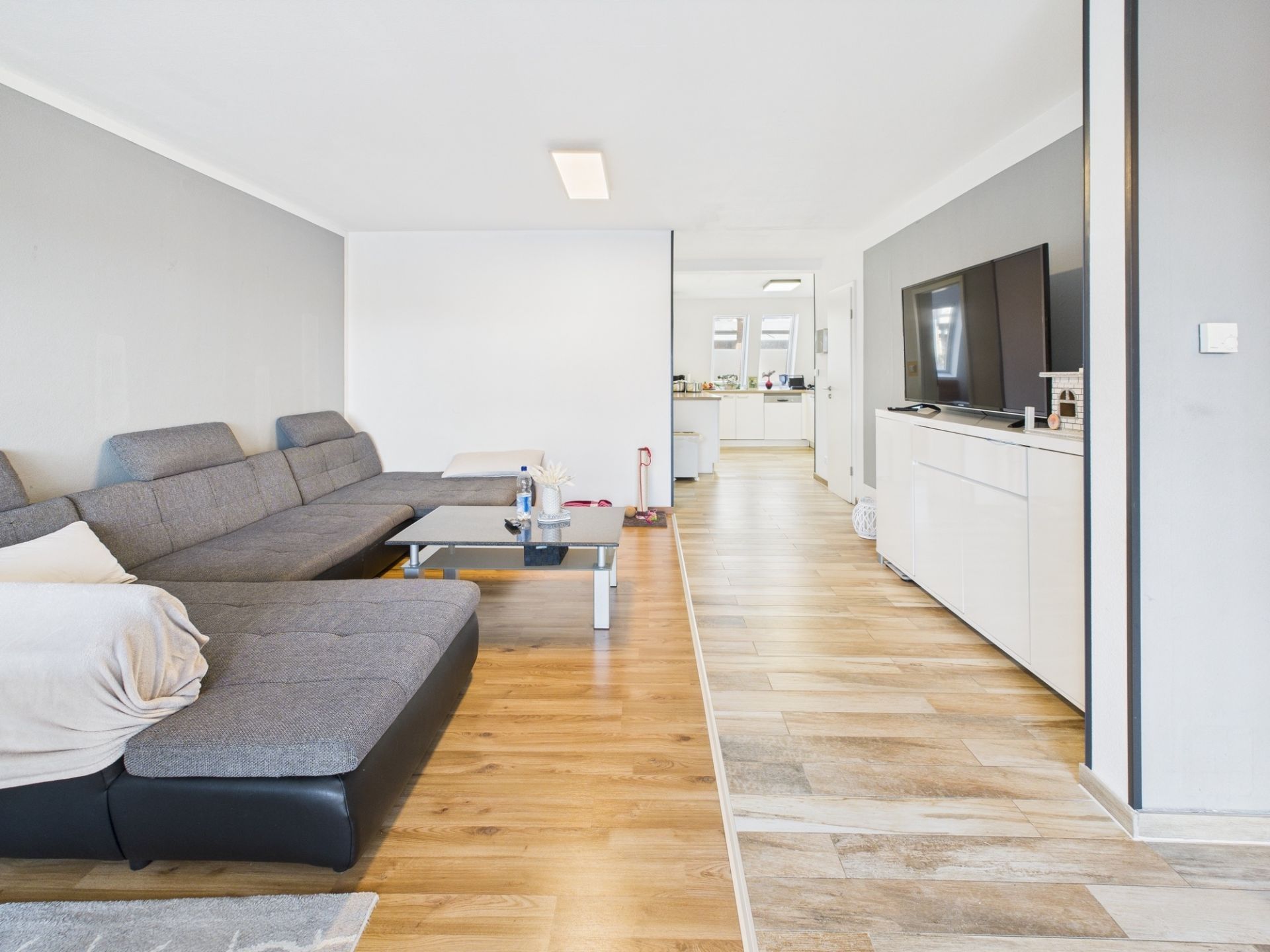 Modernes Wohnzimmer mit grauem Sofa, Couchtisch und großem Fernseher auf hellem Holzfußboden.