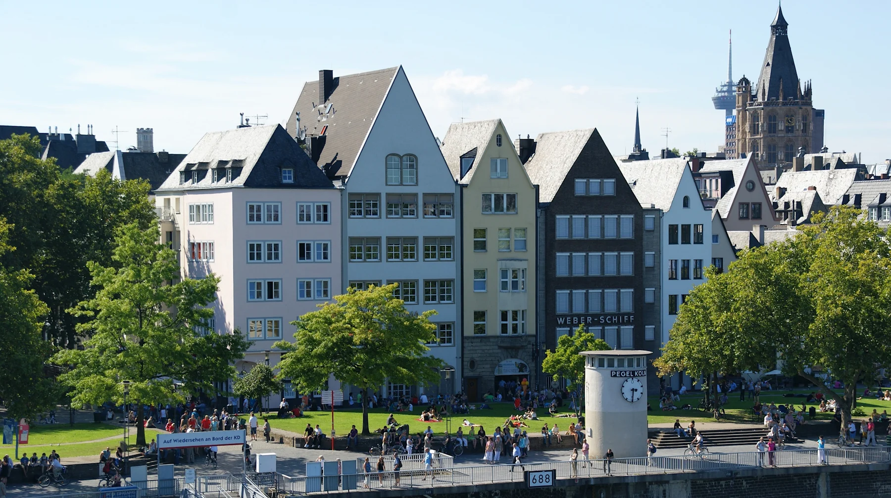 Mehrstöckige Wohnhäuser mit Satteldächern an einer grünen Uferpromenade bei sonnigem Wetter.