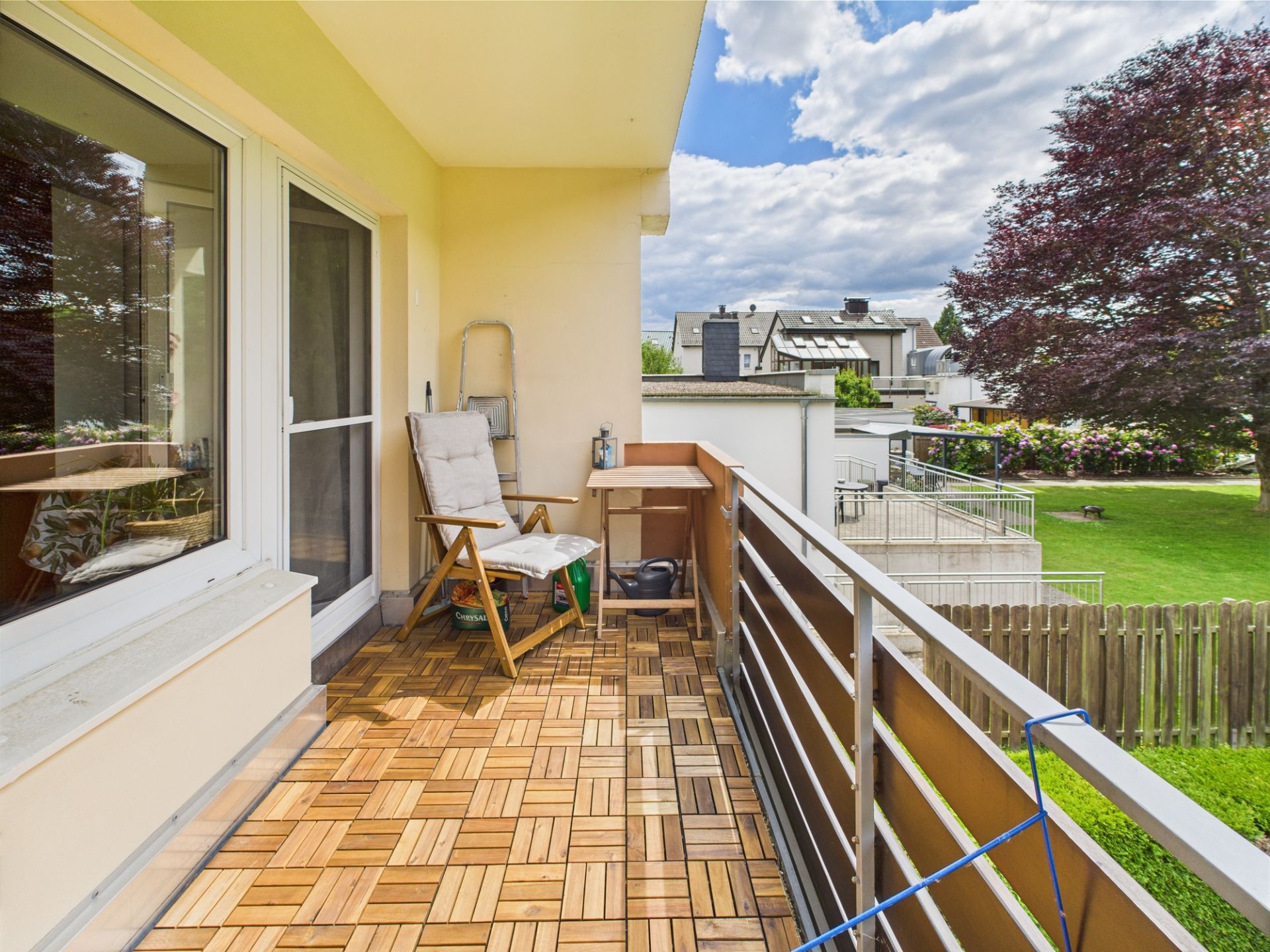 Balkon mit Holzfliesen, Stuhl und kleinem Tisch, Blick auf Garten und Bäume.