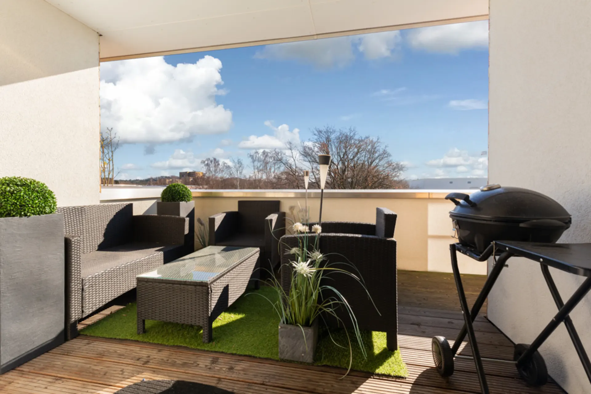 Möblierter Balkon mit Korbstühlen, Tisch, Pflanzen und Grill, Blick auf Bäume und Himmel.