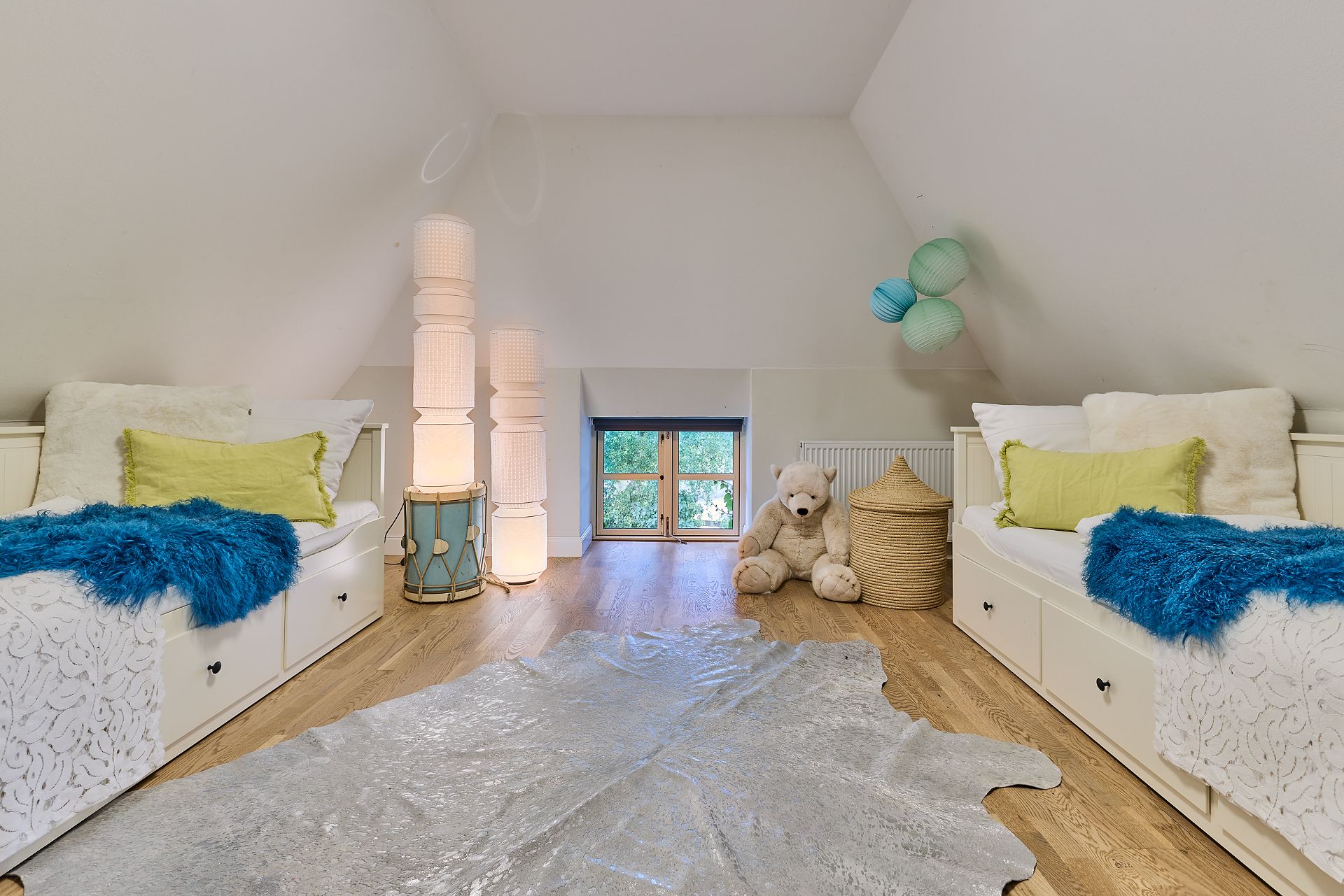 Kinderzimmer mit zwei Betten, blauem Teppich, großem Stoffbären und kleinen Fenstern.