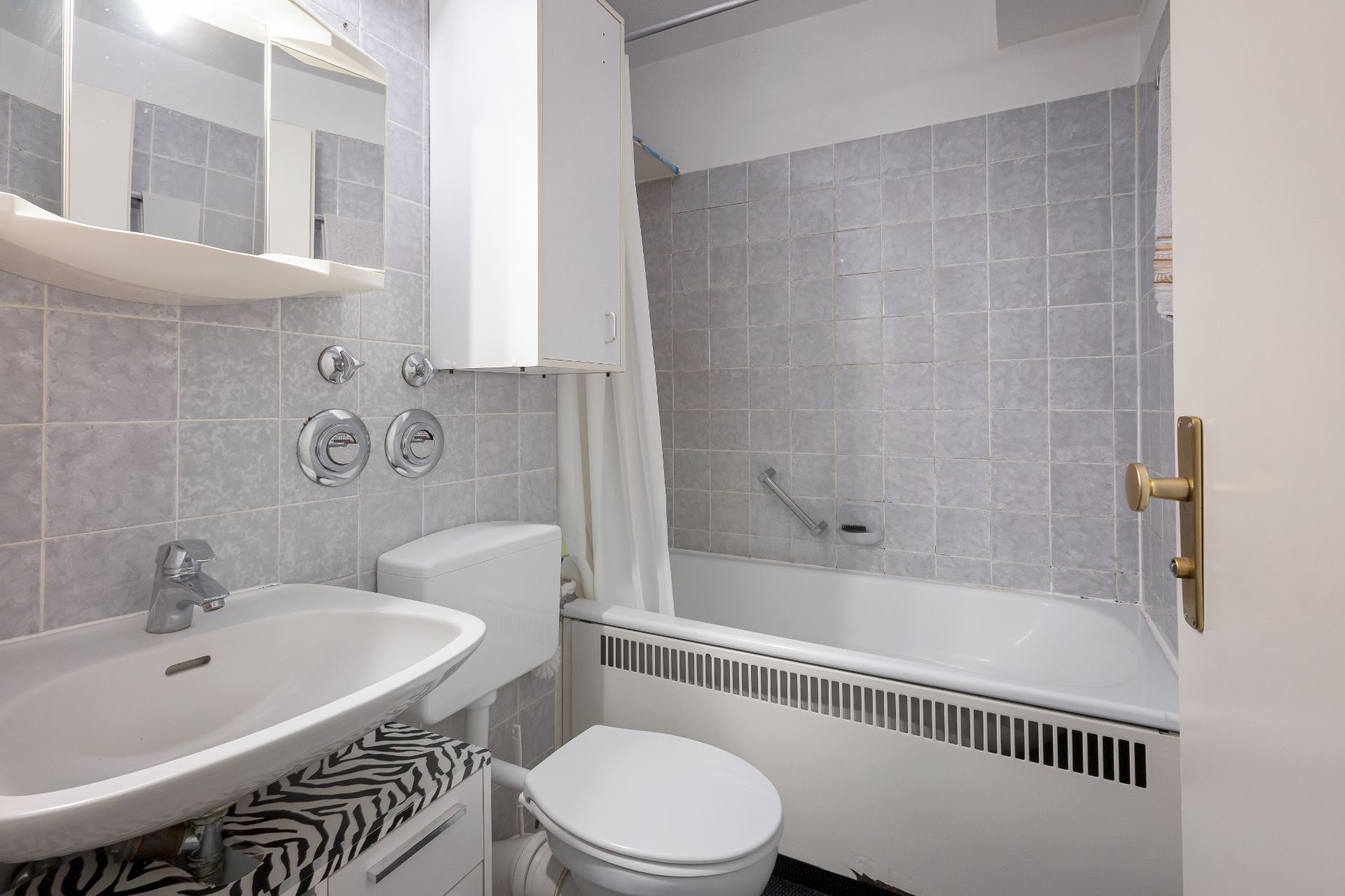 Kleines Badezimmer mit Waschbecken, WC, Badewanne und hellgrauen Wandfliesen.