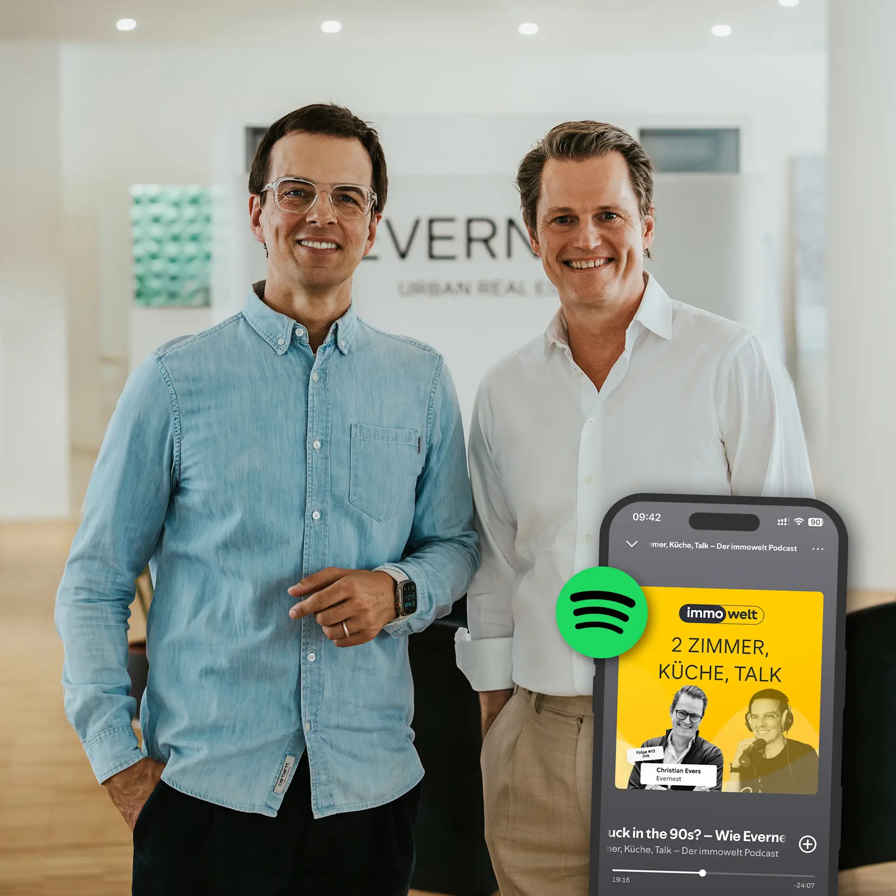 Zwei Männer stehen in einem modernen, hellen Raum. Im Vordergrund ist ein Smartphone mit Podcast-Anzeige zu sehen.