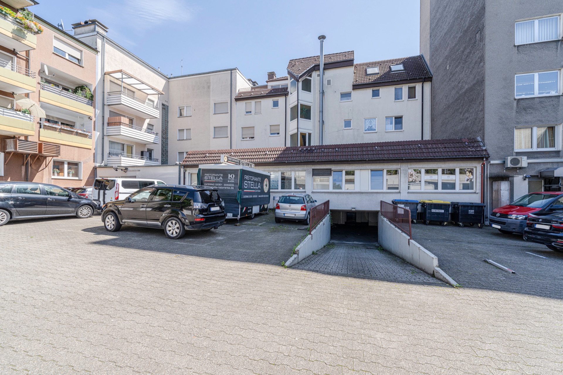 Wohnhaus mit mehreren Autos und Mülltonnen auf einem gepflasterten Hof, Zufahrt zur Tiefgarage sichtbar.