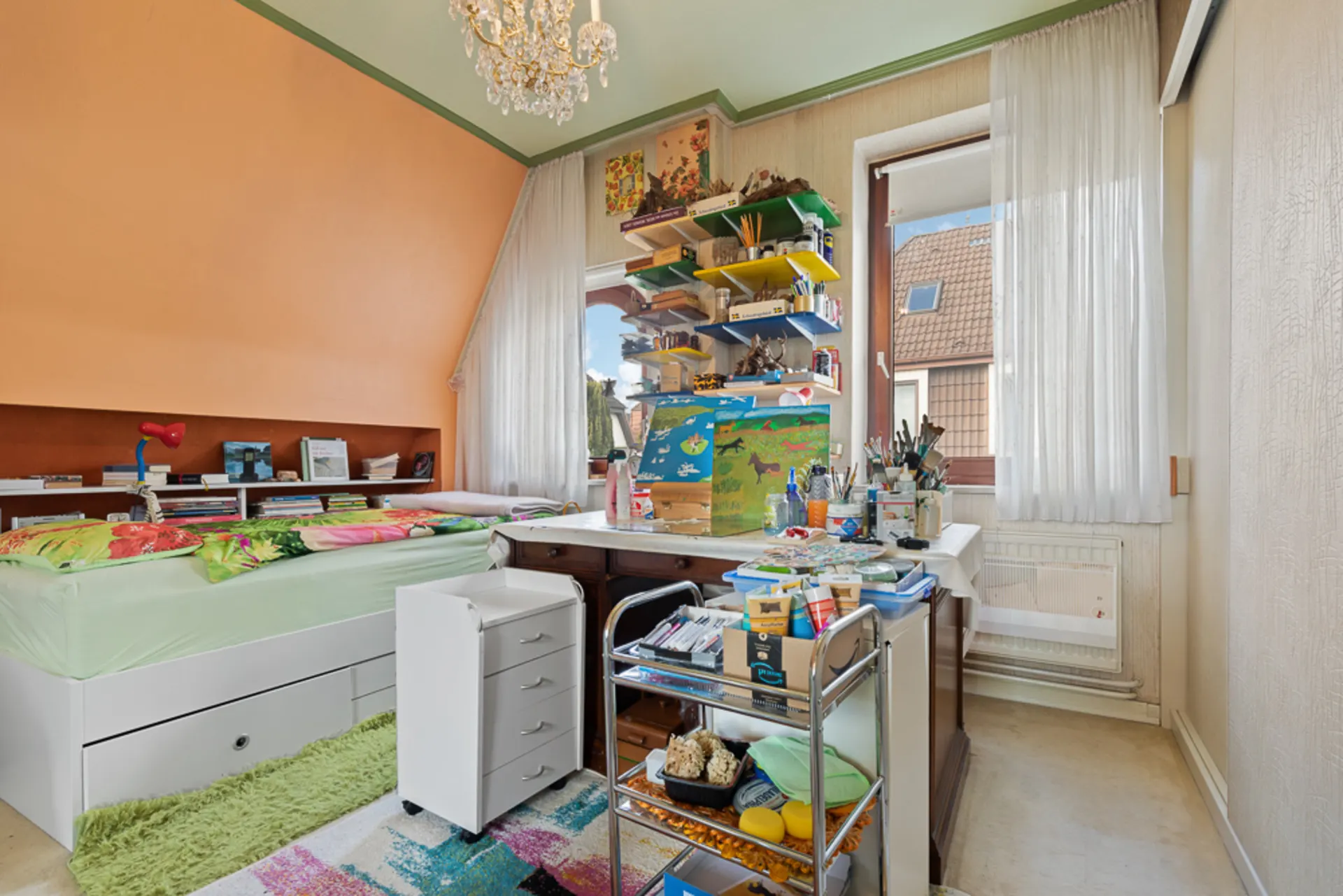 Helles Kinderzimmer mit Bett, Schreibtisch, Regalen und Bastelmaterialien vor zwei Fenstern.
