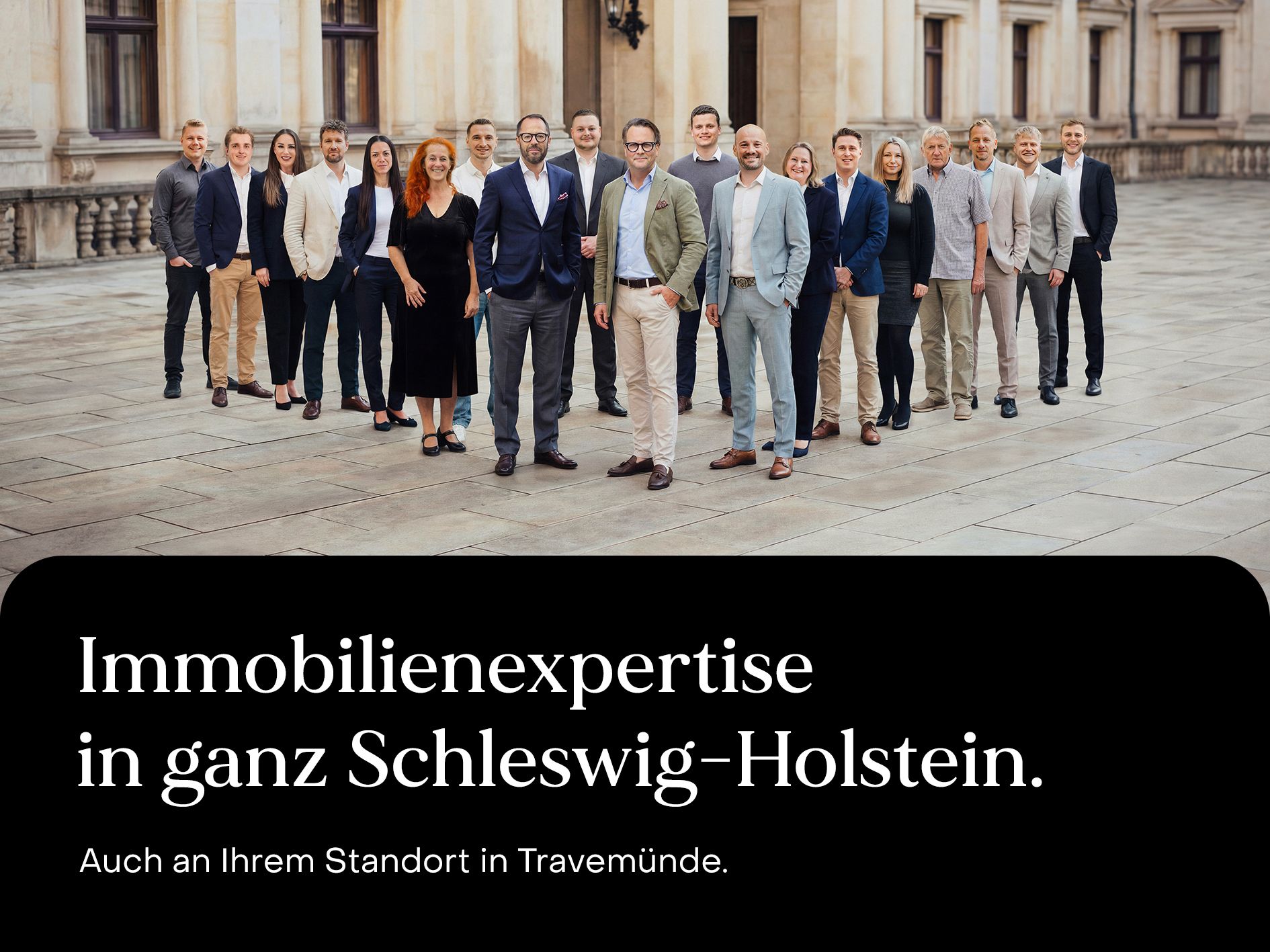 Eine Gruppe von etwa zwanzig Personen steht auf einem gepflasterten Platz vor einem großen Gebäude.