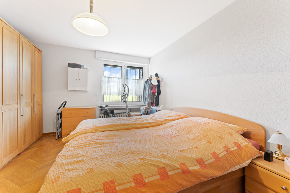 Schlafzimmer mit Doppelbett, Holzschränken, Kommode und Fitnessgerät vor Fenstern.