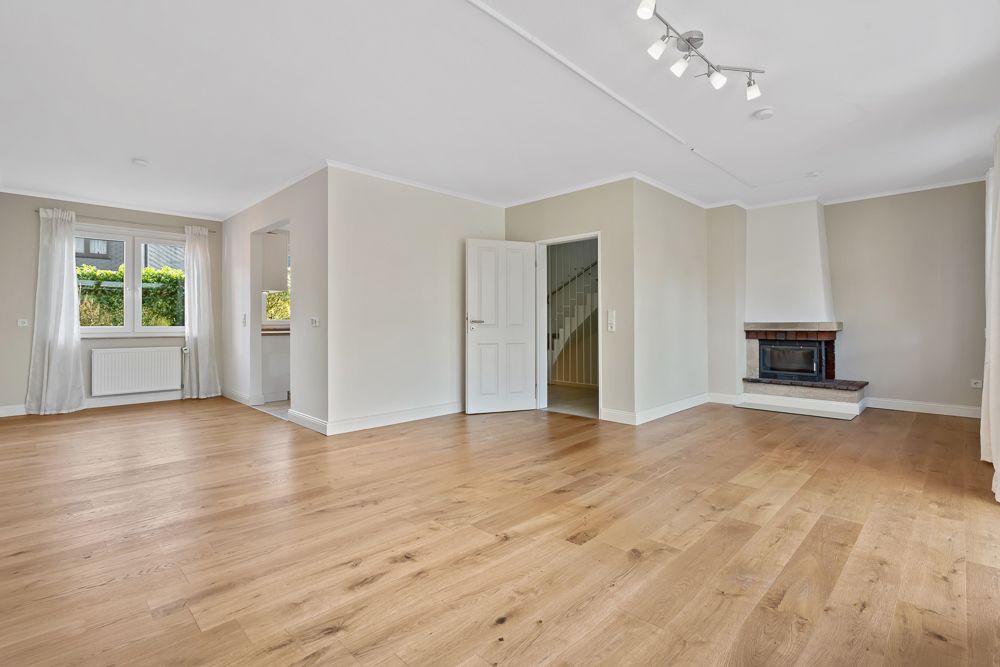 Helles Wohnzimmer mit Holzboden, Kamin, Fenstern mit weißen Vorhängen und offener Tür zu Treppe.