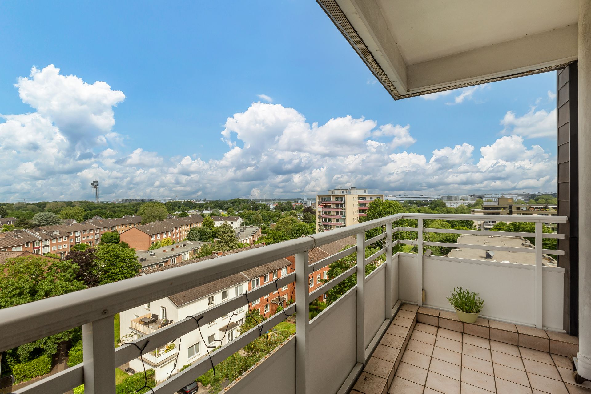 Balkon mit Fliesenboden, Blick auf Wohnhäuser, Bäume und einen blauen Himmel mit Wolken.